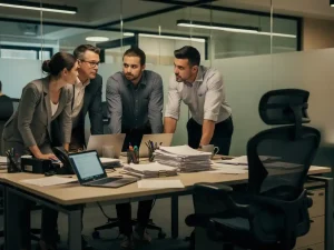 Vijf collega's rond een bureau in een open kantoor, met een opvallend lege stoel en onafgemaakte taken, sfeer van stille spanning.