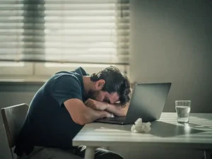 Uitgeputte thuiswerker met hoofd op gevouwen armen naast laptop, verfrommeld tissue en glas water, zacht ochtendlicht door halfgesloten jaloezieen.