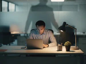 Medewerker zit gespannen en neergeslagen aan modern bureau, overschaduwd door donkere schaduw in klinisch kantoor met koel blauwgrijs licht.